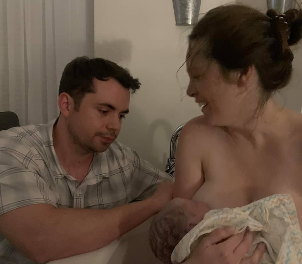 Hypno-mom sitting in tub holding newborn with partner sitting next to her