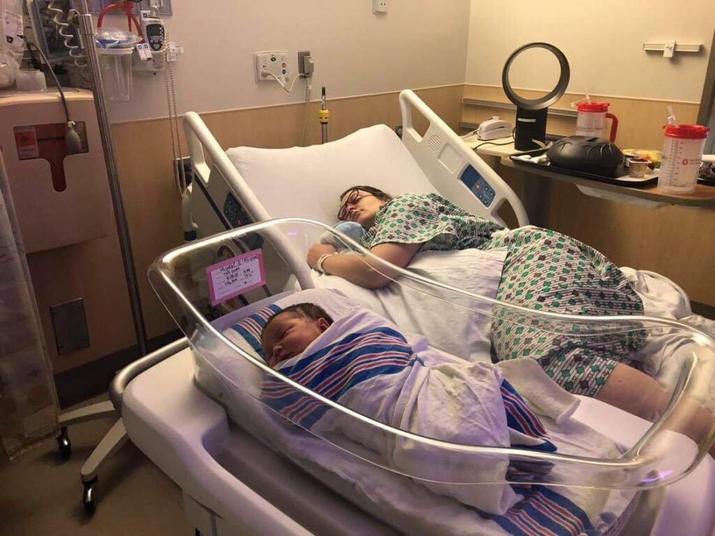 New parent sleeping in hospital bed with new baby sleeping nearby in bassinet