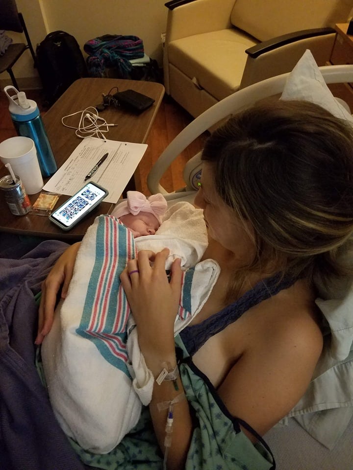 Amy in hospital bed holding her baby girl who is wrapped in a blanket with a pink hat and bow.