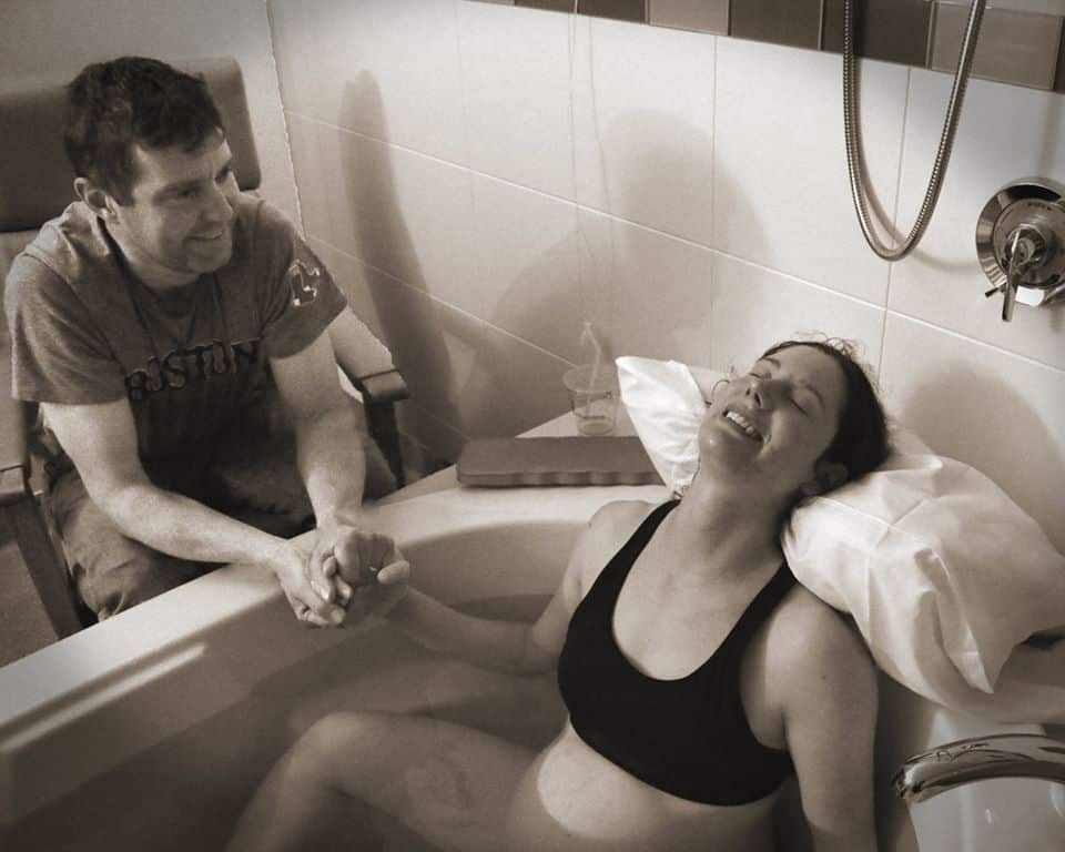 Barrett Phillips leaning back in birth tub with a smile on her face and birth partner holding her hand.
