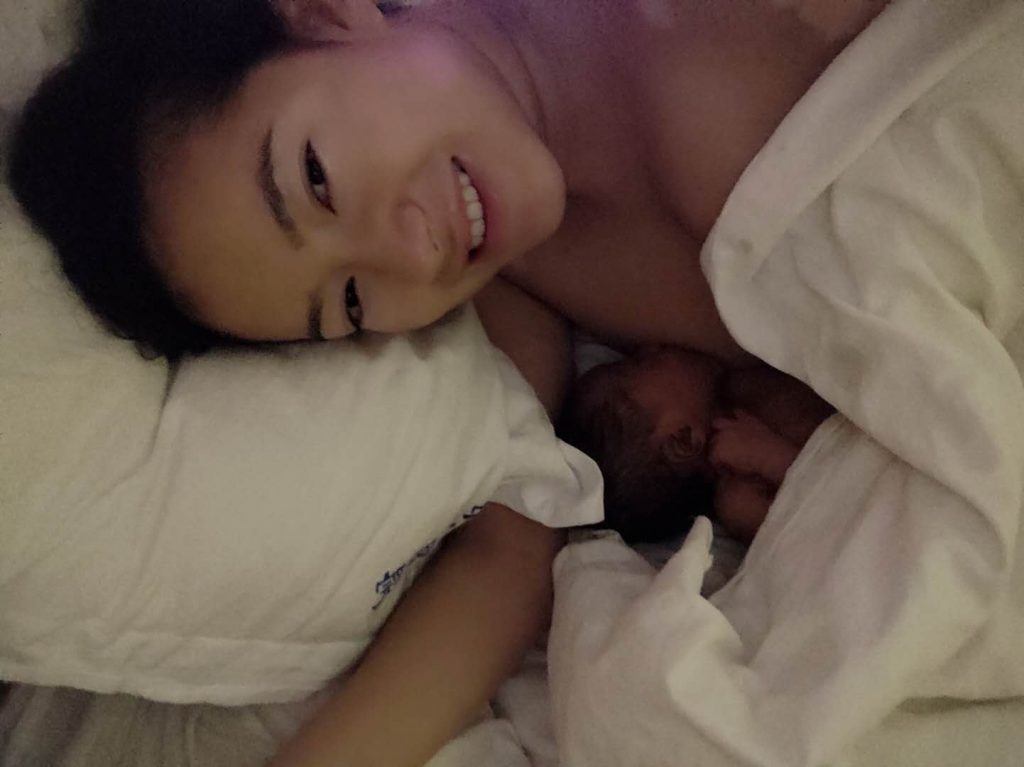 hypno-mom smiling up at camera while in bed nursing her newborn