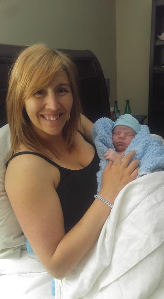 Hypno-mom Evelyne sitting in bed and smiling, holding newborn baby boy wrapped in fuzzy blue blanket and blue hat.