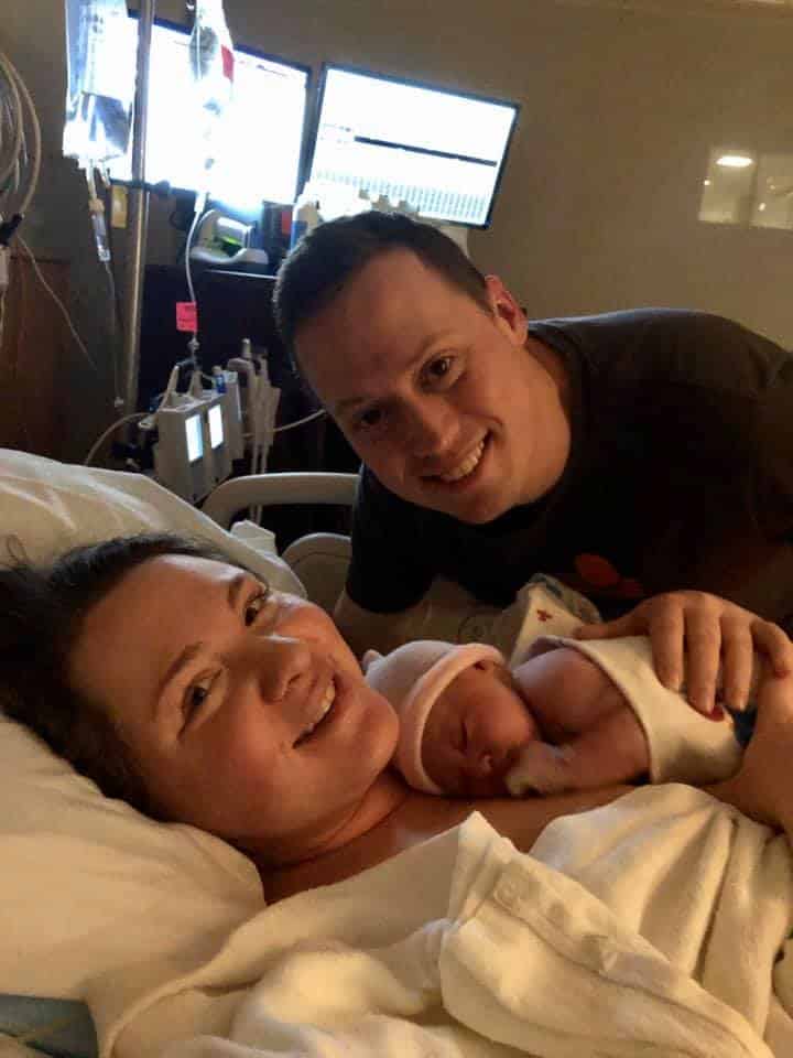 Hypno-mom Kalee, birth partner and newborn in hospital bed smiling with monitors in the background.