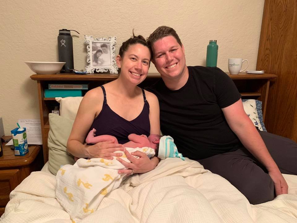 Hypno-mom Kristen and her partner sitting in their bed just after birth smiling and holding their newborn.