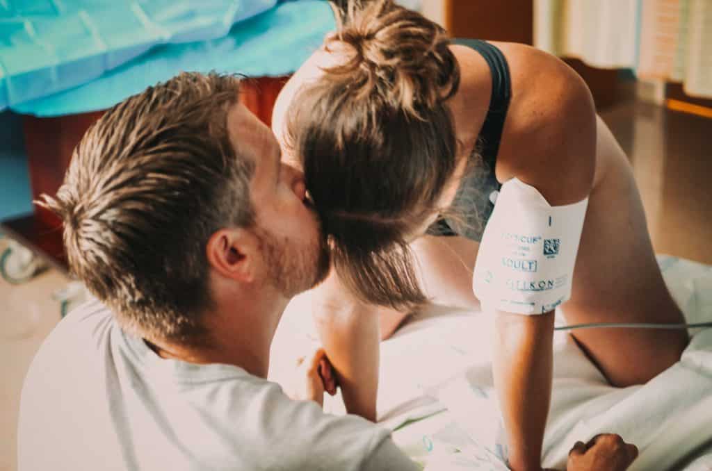 Hypno-mom Mallory kneeling on hospital bed with a sports bra on and a blood pressure cuff getting a kiss on her forehead from her partner.