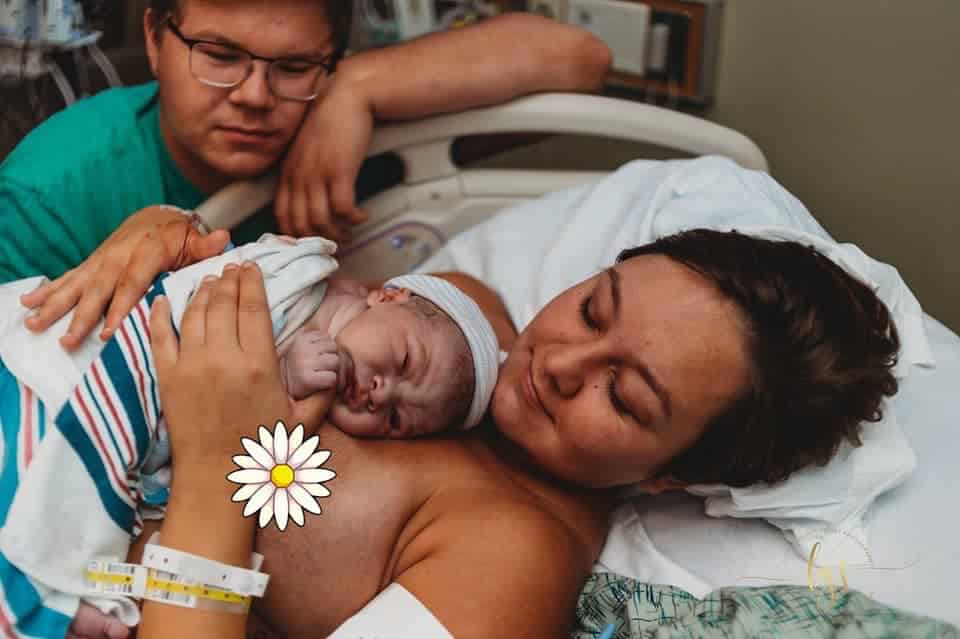 Hypno-mom morgan laying in hospital bed skin to skin with newborn baby wearing a had and covered in a blanket with partner sitting next to her resting his check on his hand.