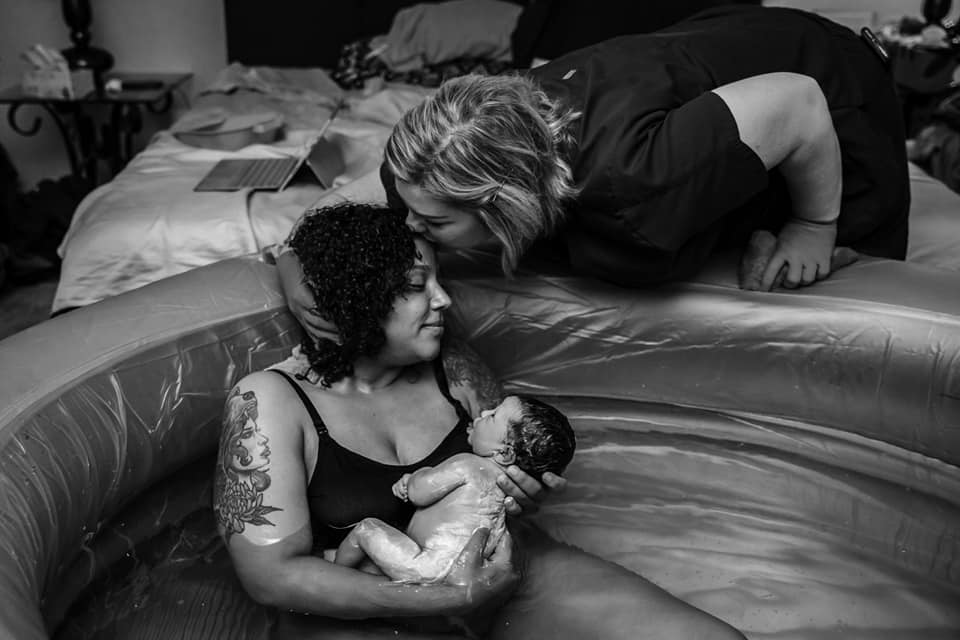 Hypno-mom Nichole being kissed on the forehead by her midwife while sitting in the tub holding her newborn baby who is covered in Vernix.