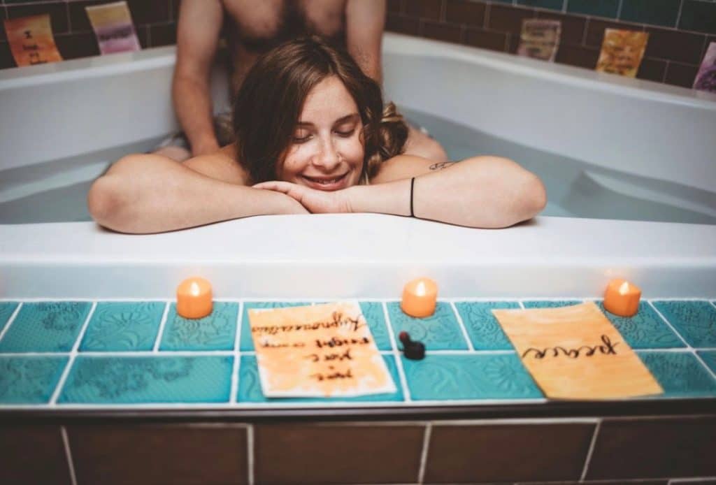 Pregnant woman relaxing in a birthing tub with her partner behind her, smiling with her eyes closed as candles and affirmation cards sit along the edge of the bath.