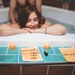 Pregnant woman relaxing in a birthing tub with her partner behind her, smiling with her eyes closed as candles and affirmation cards sit along the edge of the bath.