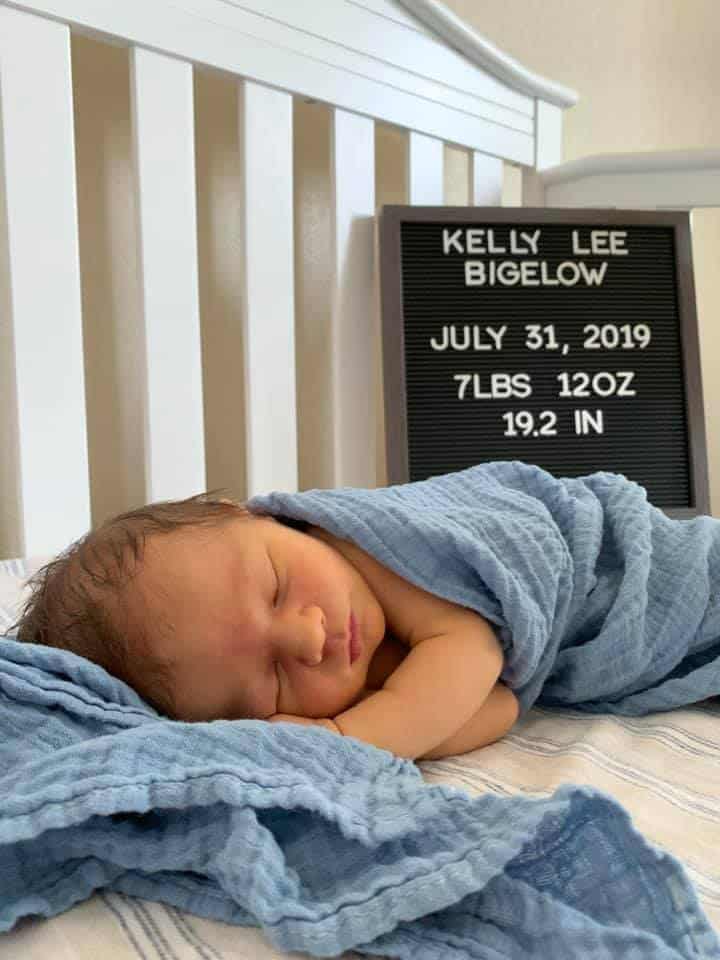 Hypno-baby Kelly wrapped in a blue blanket in a crib with a sign saying Kelly Lee Bigelow July 31, 2019 7lbs 12 oz 19.2 in.